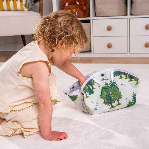 A young child in a cream dress kneels on a rug and reaches into a forest-patterned storage basket beside the TP Toys Gruffalo Play Set with Wooden Characters, displayed on a double-sided fabric playmat, in a cozy, organized room.