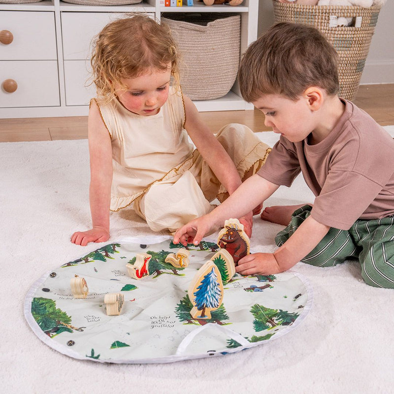Two young children sit on a white carpet, playing with the TP Toys The Gruffalo Play Set with Wooden Characters on a round, double-sided fabric playmat in a tidy room.