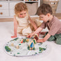 Two young children sit on a white carpet, playing with the TP Toys The Gruffalo Play Set with Wooden Characters on a round, double-sided fabric playmat in a tidy room.