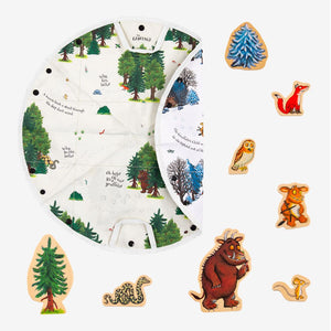 A circular illustrated playmat featuring a forest scene is partially folded open and surrounded by TP Toys’ The Gruffalo Play Set with Wooden Characters, including charming figures such as a fox, owl, mouse, snake, Gruffalo, and trees. This delightful double-sided fabric playmat perfectly complements the set and inspires imaginative woodland adventures.