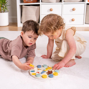 Two young children play with The Gruffalo Wooden Counting Clock by TP Toys, an FSC® certified numeracy puzzle, on a white carpet in a bright, organized room with storage baskets and a plant in the background.