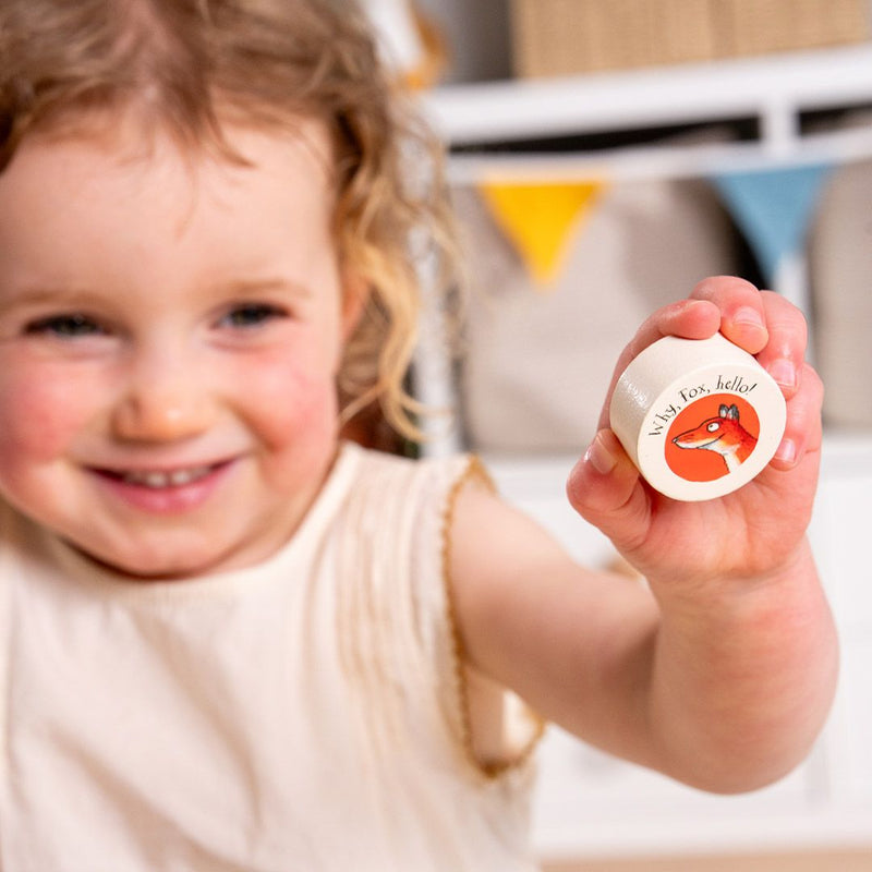 A young child holds up a small round container featuring a fox illustration and the text “Hey, fox, hello,” while nearby sits the TP Toys The Gruffalo Wooden Counting Clock – FSC® certified: a charming counting toy ideal for supporting early numeracy skills.