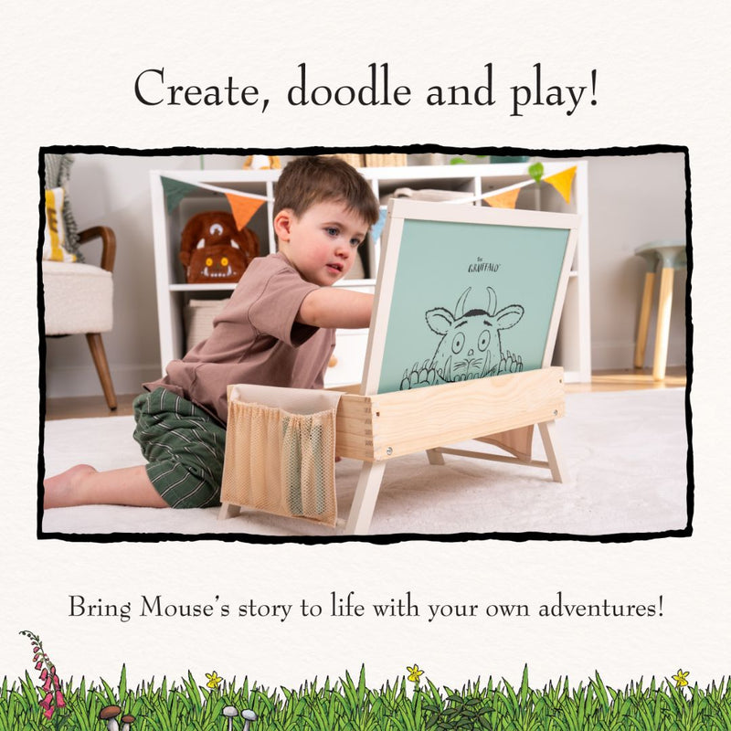 A young boy uses the TP Toys Gruffalo Wooden Creative Station - FSC® certified art easel to draw a cartoon mouse in a playroom, surrounded by toys, books, and magnetic pieces.