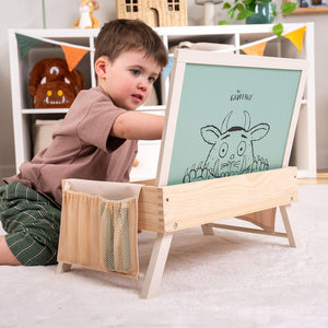 A young child sits on the floor drawing at The Gruffalo Wooden Creative Station – FSC® certified by TP Toys, which features a Gruffalo illustration. The creative station is set in a playroom filled with toys, decorations, and magnetic pieces in the background.