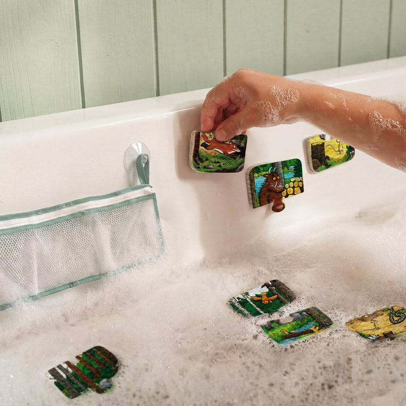A child’s hand places TP Toys The Gruffalo Foam Bath Figures—colorful animal-shaped foam pieces—on a white bathtub wall above sudsy bathwater, next to a mesh storage pouch.