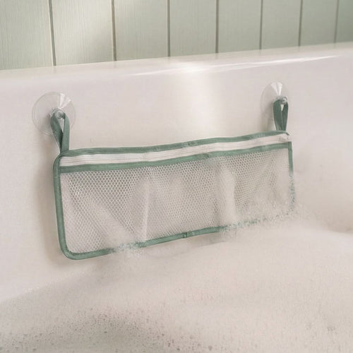 A mesh storage pouch with green edges from TP Toys is attached to a tiled wall above a bathtub using two suction cups. Partially covered by bubbles and filled with The Gruffalo Foam Bath Figures, it keeps bath time toys organized and within easy reach.