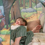A child in striped pajamas lies on a blanket inside the TP Toys The Gruffalo Play Tent, surrounded by illustrated forest scenes, smiling and gazing upward, immersed in imaginative play.