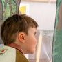 A young boy with short brown hair peers through the mesh screen of the TP Toys The Gruffalo Play Tent, surrounded by illustrated trees, with soft indoor lighting in the background.