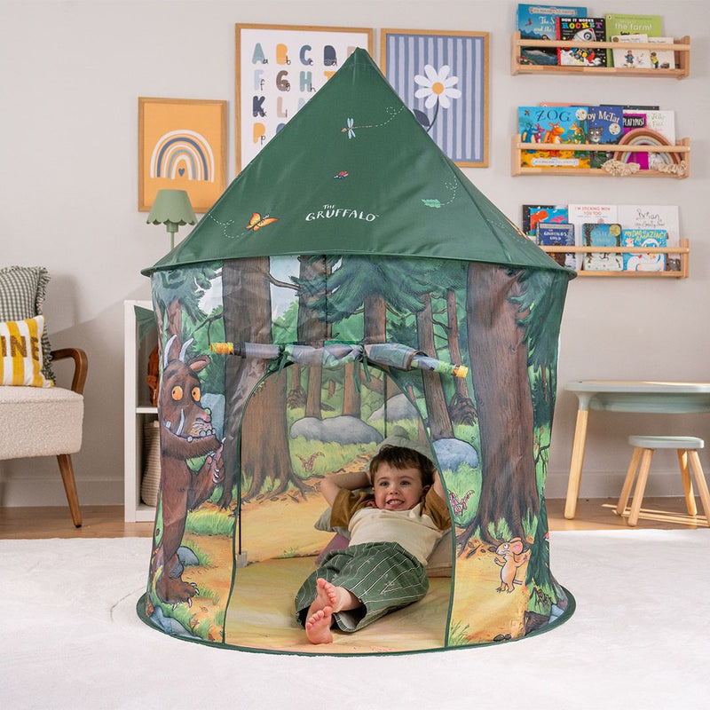 A young child lounges inside the TP Toys The Gruffalo Play Tent, adorned with delightful illustrations from The Gruffalo, in a cozy, book-filled playroom—perfect for imaginative play.