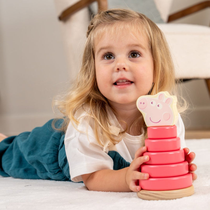 A young girl lies on the floor holding a pink stacking toy with a cartoon pig face from the TP Toys Grow with Peppa Pig Wooden Roller Rattle and Topple Tower Set.