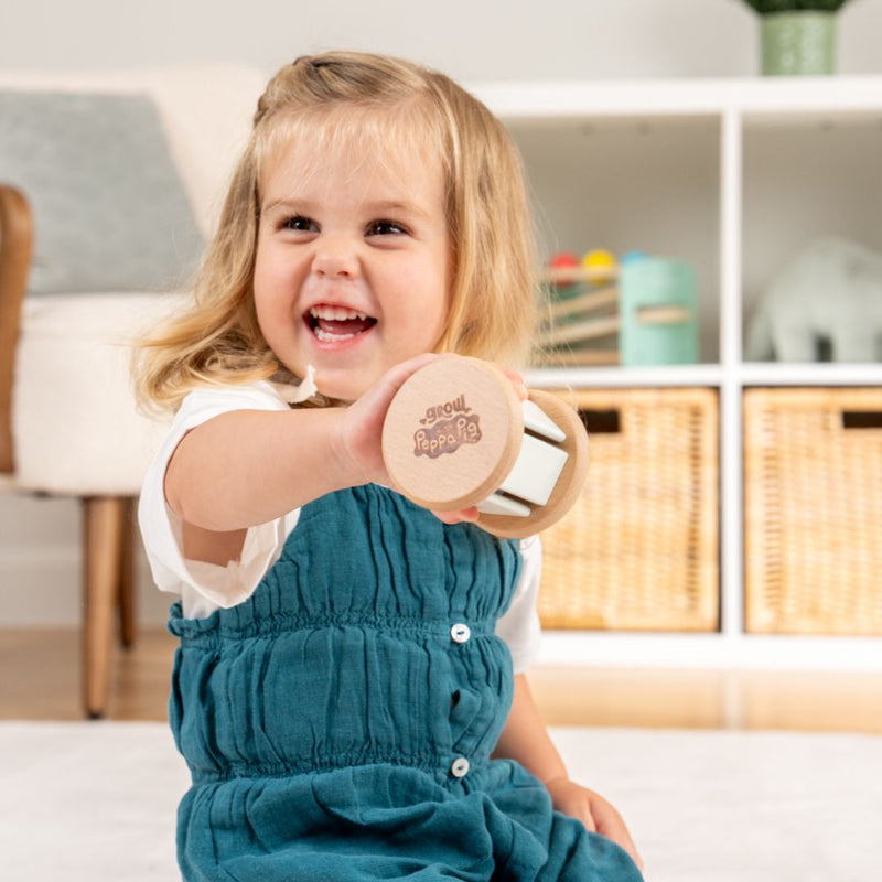 A young child in a teal dress smiles and holds up the TP Toys Grow with Peppa Pig Wooden Roller Rattle and Topple Tower Set indoors, with shelves and baskets in the background.