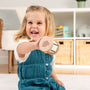 A young child in a teal dress smiles and holds up the TP Toys Grow with Peppa Pig Wooden Roller Rattle and Topple Tower Set indoors, with shelves and baskets in the background.