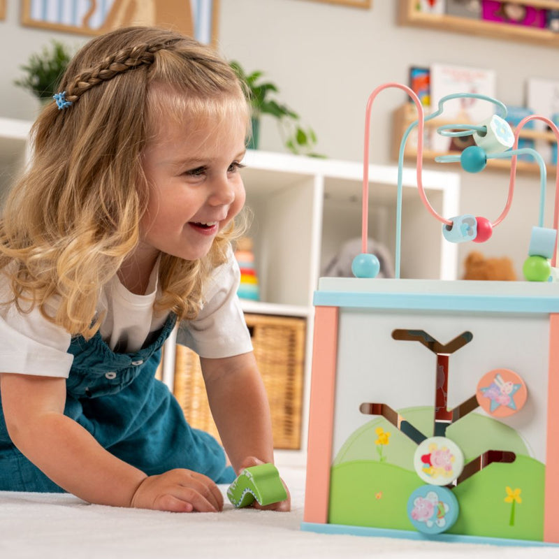 A young blonde girl explores sensory play on the floor with the TP Toys Grow with Peppa Pig Wooden Activity Cube, using its colorful bead and shape puzzles to develop her fine motor skills.