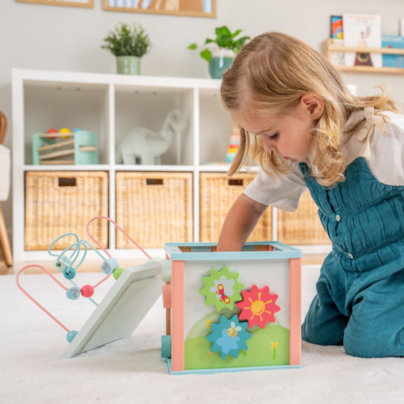 A young child in blue overalls explores and develops fine motor skills with the TP Toys Grow with Peppa Pig Wooden Activity Cube on a white rug in a bright, organized playroom.