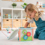 A young child in blue overalls explores and develops fine motor skills with the TP Toys Grow with Peppa Pig Wooden Activity Cube on a white rug in a bright, organized playroom.