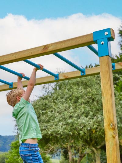 A child hangs from blue monkey bars on the TP Toys Skywood Wooden Climbing Frame, featuring extra play towers, slides, swings, and more—perfect for outdoor fun. FSC® certified.