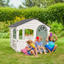 Three children sit on the grass by the TP Toys Palplay Plastic Playhouse of Fun Snowdrift, enjoying outdoor role play and sharing colorful cups together in the garden.