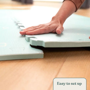 A hand assembles TP Toys’ TP Active-Tots Premium Play Safety Mats in light blue non-toxic foam on a wooden surface, with an “Easy to set up” label in the corner.