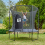 A child enjoys bouncing on the TP Toys TP Genius® 8ft Round Trampoline with safety netting, set on grass among trees and bushes, highlighting the safe design of this trampoline.