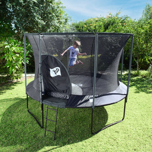 A child jumps on a TP Toys TP Genius® 10ft Round Trampoline with SurroundSafe enclosure and galvanised steel frame in a grassy backyard surrounded by trees and bushes.