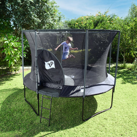 A child jumps on a TP Toys TP Genius® 10ft Round Trampoline with SurroundSafe enclosure and galvanised steel frame in a grassy backyard surrounded by trees and bushes.