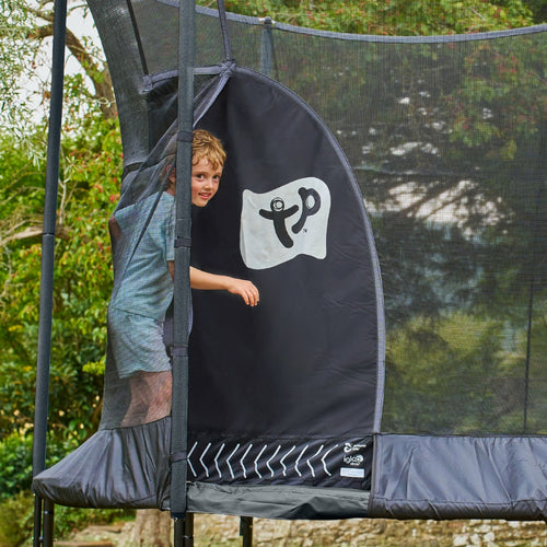 A young boy in casual clothes enters a TP Toys TP Genius® 10ft Round Trampoline with a galvanised steel frame and SurroundSafe enclosure, set up outdoors with trees in the background.