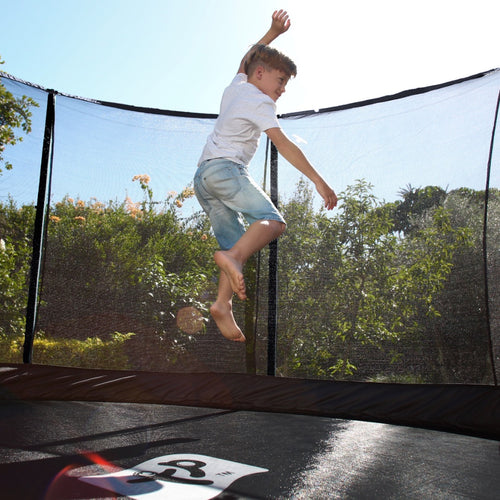 A young boy in a white shirt and denim shorts jumps on a TP Toys TP Genius® 10ft Round Trampoline with a robust galvanised steel frame and SurroundSafe enclosure, set outdoors among trees on a sunny day.