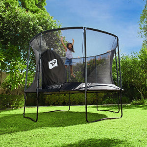 A child is jumping on a TP Toys Genius® 12ft Round Trampoline with safety enclosure and galvanised steel frame in a grassy backyard among trees and bushes.