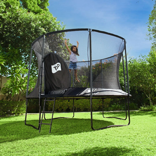 A child jumps on a TP Toys TP Genius® 12ft Round Trampoline with SurroundSafe enclosure and galvanized steel frame in a grassy backyard surrounded by trees and bushes.