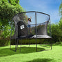 A child jumps on a TP Toys TP Genius® 12ft Round Trampoline with SurroundSafe enclosure and galvanized steel frame in a grassy backyard surrounded by trees and bushes.