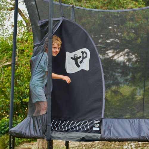 A child steps out of a TP Genius® 12ft Round Trampoline by TP Toys, holding the entrance flap of the SurroundSafe enclosure open. The galvanised steel frame gleams with trees and greenery visible in the background.