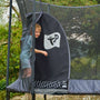A child steps out of a TP Genius® 12ft Round Trampoline by TP Toys, holding the entrance flap of the SurroundSafe enclosure open. The galvanised steel frame gleams with trees and greenery visible in the background.