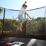 A boy in a white shirt and denim shorts is jumping outdoors on a TP Toys TP Genius® 12ft Round Trampoline, featuring a SurroundSafe enclosure, greenery, and a strong galvanised steel frame.