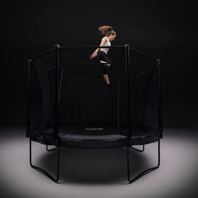 A young girl in athletic wear jumps high on the TP Toys 12ft Infinity Round Trampoline with safety netting, set against a dark background, highlighting its superior bounce and outstanding safety features.