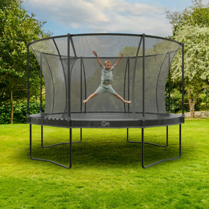 A child jumps with arms and legs spread on the TP Toys 14 ft Infinity Round Trampoline, featuring superior bounce and safety, set on a green lawn with trees in the background.