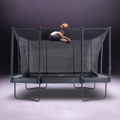 A person does a tuck jump high above the TP Toys Infinity 10ft x 14ft Rectangular Trampoline with net enclosure, set against a neutral background. Easy assembly adds convenience.