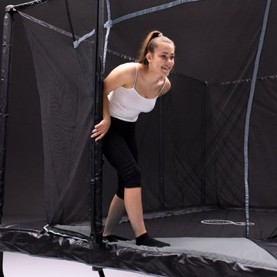 A young woman in athletic wear enters the TP Toys Infinity 10ft x 14ft Rectangular Trampoline through a zippered safety net, enjoying its easy assembly and Olympic-approved design.
