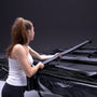 A woman in athletic wear secures a strap on the folded TP Toys Infinity 10ft x 14ft Rectangular Trampoline against a dark background, showcasing its easy assembly.