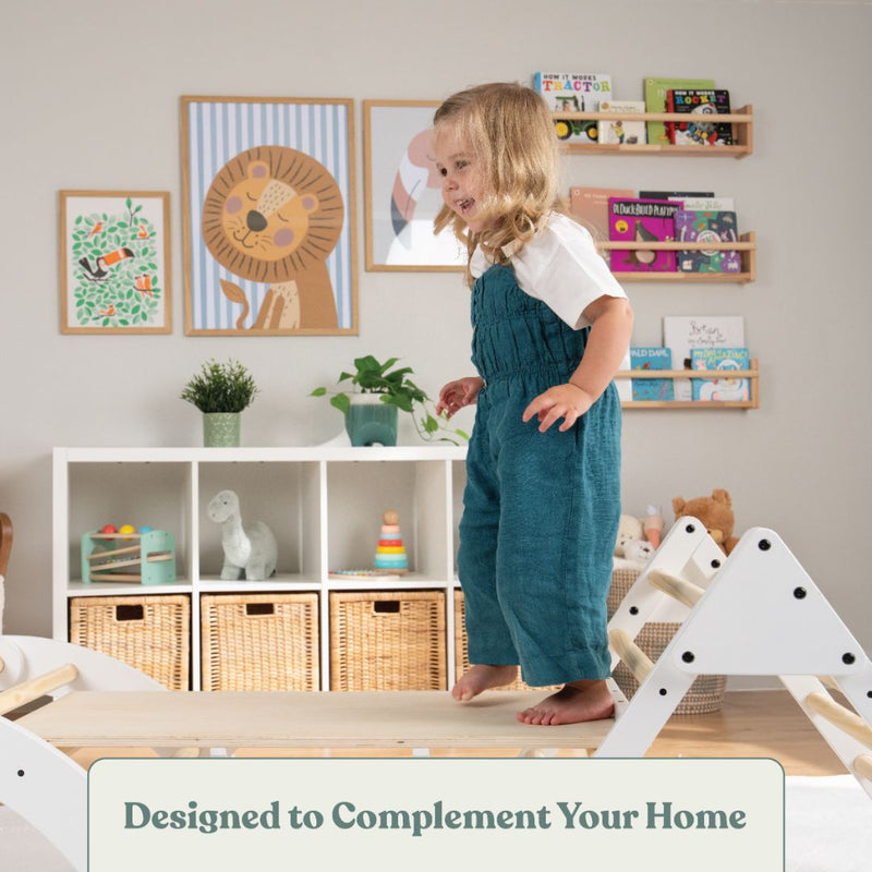 A young child stands on the TP Toys TP Active-Tots Pikler Style Three in One Playset—FSC® certified—in a playroom with animal-themed decor, toys, and bookshelves. Text reads: Designed to Complement Your Home.