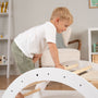 A young boy in a white shirt and green shorts climbs the TP Toys TP Active-Tots Pikler Style Three in One Playset—FSC® certified—exploring the Montessori-inspired climbing set with curiosity and joy in a bright, modern room.