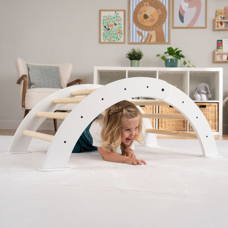 A young girl crawls under the TP Active-Tots Pikler Style Three in One Playset by TP Toys, an FSC® certified Montessori-inspired white wooden climbing arch, in a bright playroom decorated with animal art.