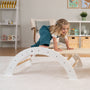 A young girl climbs over the TP Toys TP Active-Tots Pikler Style Three in One Playset - FSC® certified, in a tidy, well-lit living room with shelves, baskets, and children's decor.