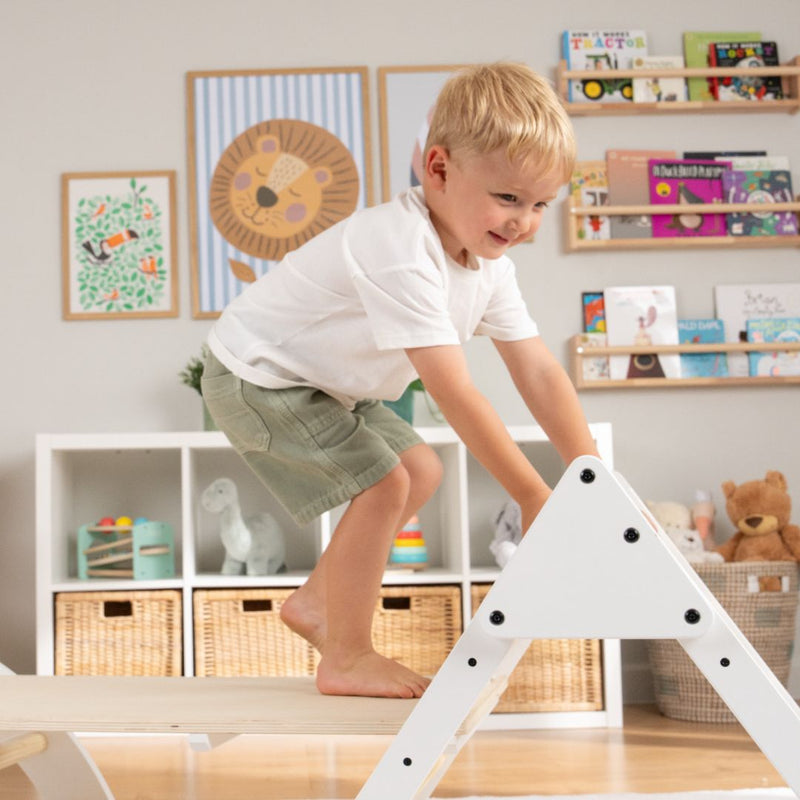 A young boy in a white shirt and green shorts climbs on the TP Active-Tots Pikler Style Three in One Playset by TP Toys in a brightly decorated children's room.