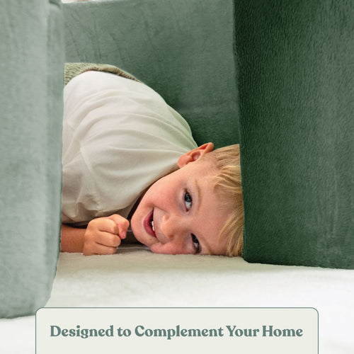 A young child smiles while lying on the floor, surrounded by the TP Toys Active-Tots Pikler Style Rainbow Arch Playset in green and gray padding. Text at the bottom reads, Designed to Complement Your Home.