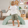A young child in a white shirt and green shorts develops gross motor skills playing with the TP Toys Active-Tots Pikler Style Rainbow Arch Playset in a bright, organized playroom equipped with Montessori toddler play equipment.