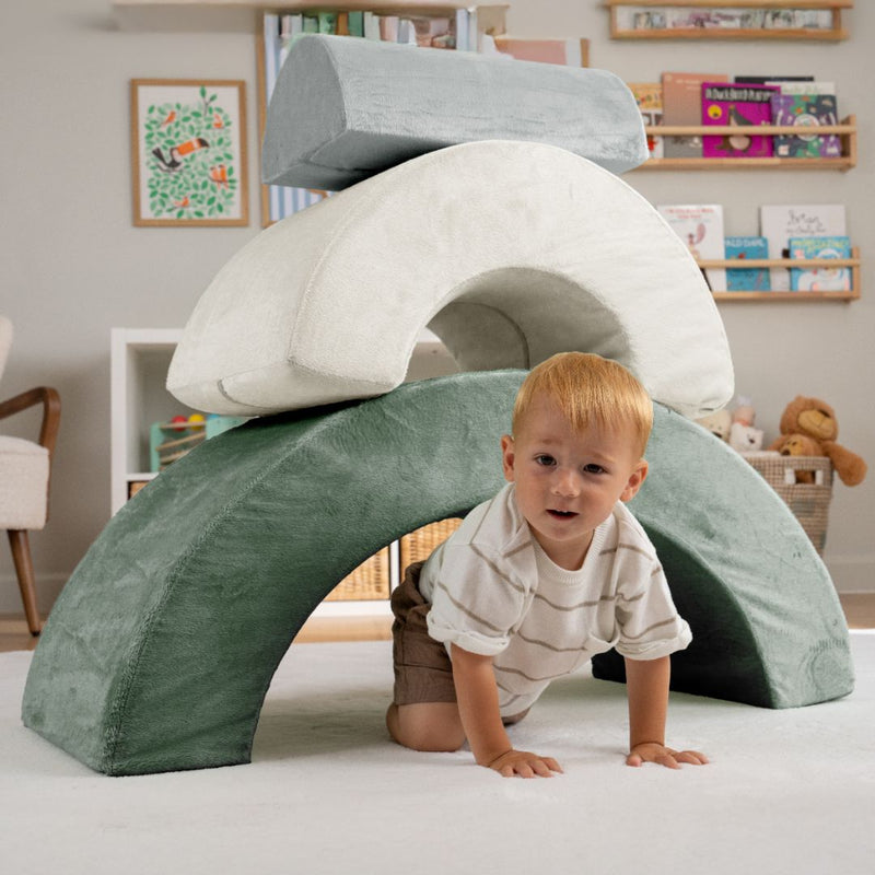 A young child crawls under the TP Toys TP Active-Tots Pikler Style Rainbow Arch Playset in a living room, building gross motor skills with this Montessori-inspired toddler play equipment.