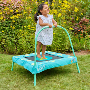 A young child in a light dress enjoys indoor or outdoor play on the TP Toys Bluey Junior 3ft Trampoline with handlebar, surrounded by green grass and yellow flowers.
