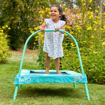 A young girl enjoys her first TP Toys Bluey Junior 3ft Trampoline with handlebar in the garden, surrounded by green grass and yellow flowers—ideal for indoor or outdoor play.