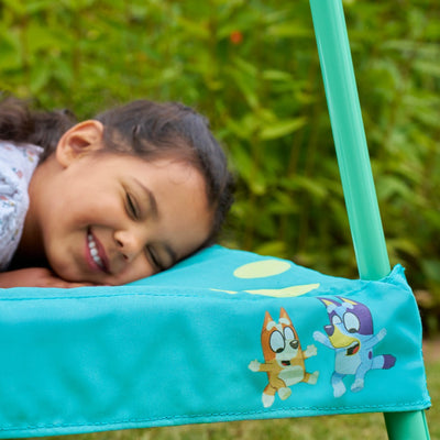 A young girl smiles, resting on a blue chair with cartoon characters, dreaming of her first TP Toys Bluey Junior 3ft Trampoline for indoor and outdoor fun, with greenery in the background.