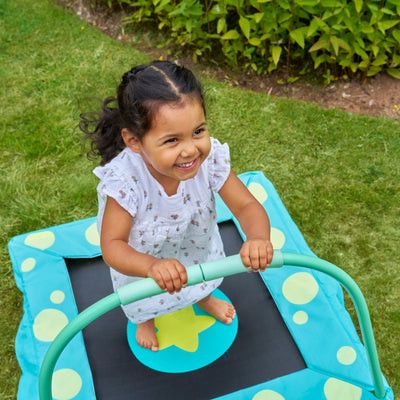 A young child in a white dress smiles while holding the handlebar of the TP Toys Bluey Junior 3ft Trampoline, enjoying their first trampoline experience outdoors with grass and green bushes in the background.